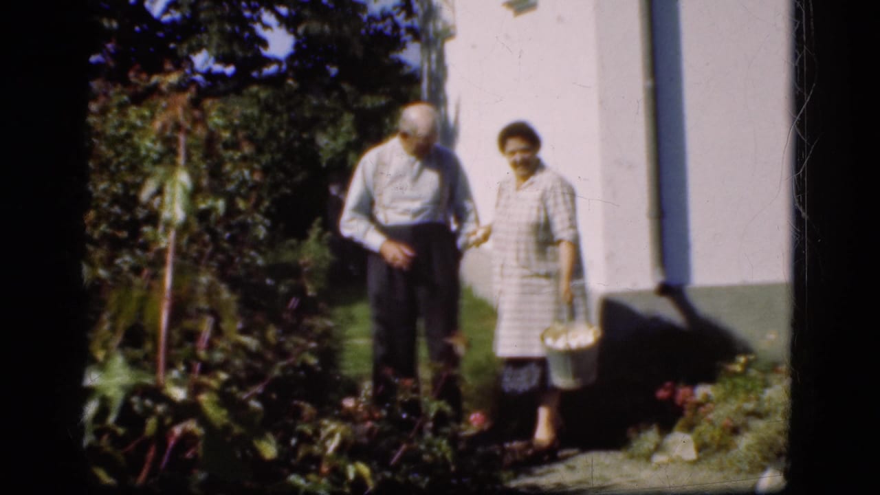 Elderly couple standing beside a white building in a garden, with the woman holding a basket.