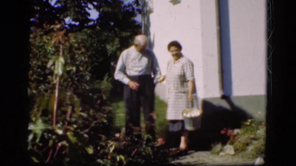 In Denmark 1975 home-movie footage, an elderly couple stands by a white garden wall while the woman holds a basket.
