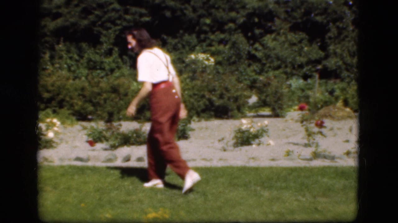 A young person walks across a Danish garden lawn in 1975 footage, with flower beds and shrubs behind them.