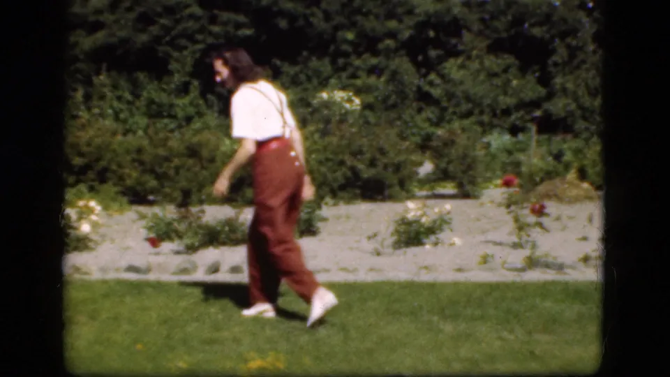 A young person walks across a Danish garden lawn in 1975 footage, with flower beds and shrubs behind them.