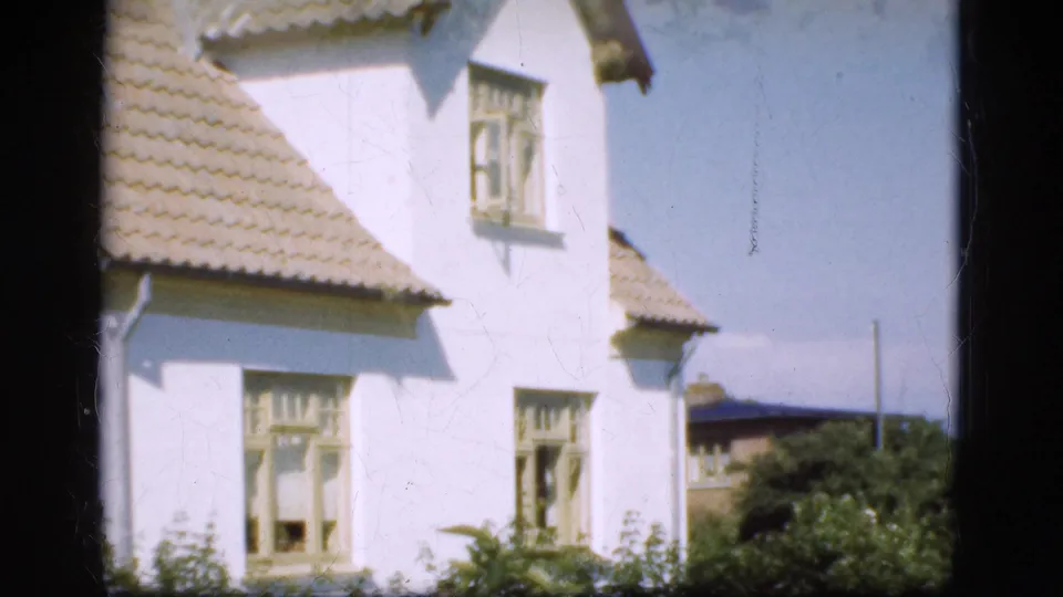 1975 Denmark home-movie frame of a white two-story house with tiled roof and pale window trim, seen through dark vertical framing.