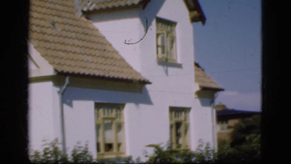 1975 Denmark home-movie frame showing a closer view of a white residential exterior with roof tiles, windows, and shrubs.