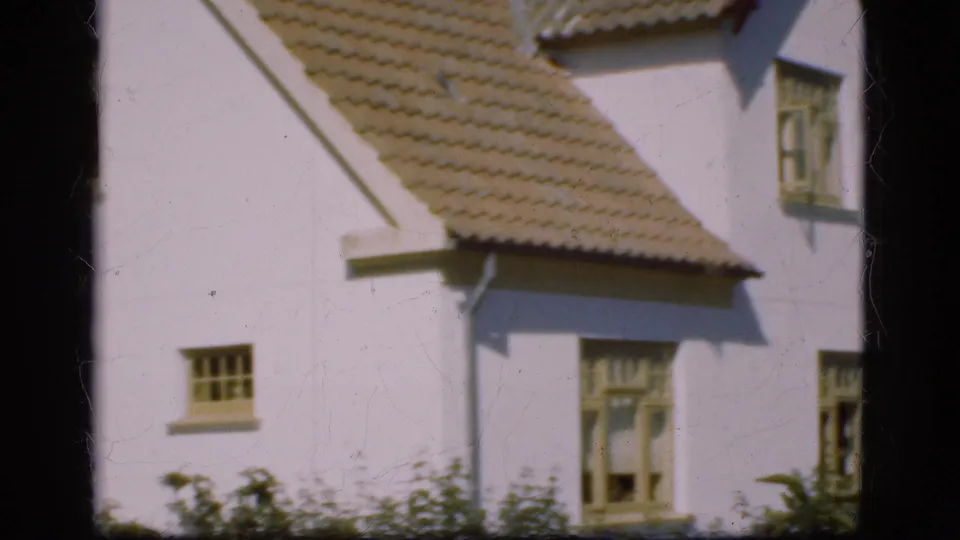 1975 Denmark home-movie frame of a white house gable and tiled roof, with small windows and garden shrubs in front.