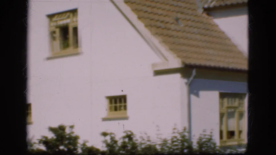 1975 Denmark home-movie frame showing a white house exterior with tiled roof, windows, grass, shrubs, and dark film edges.