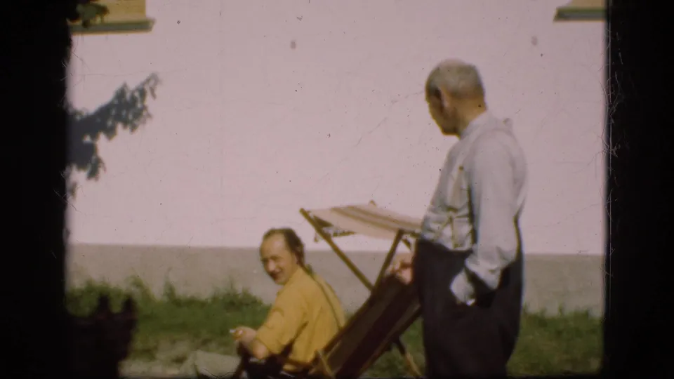 1975 Denmark home-movie frame of two adult men outside, one seated and looking toward the camera while the other stands near the chair.