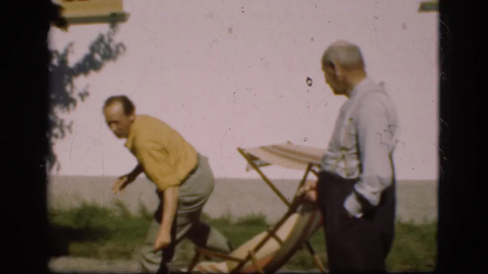 1975 Denmark 8mm frame showing a man rising from a folding deck chair as another adult man stands nearby outside the white house.