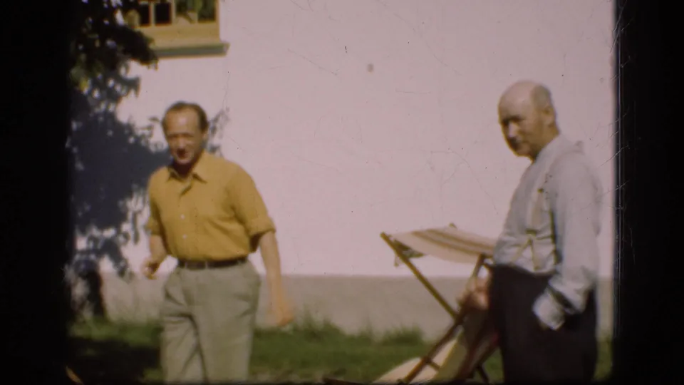 In Denmark 1975 home-movie footage, two adult men stand outdoors on grass near wooden folding chairs and a white wall.