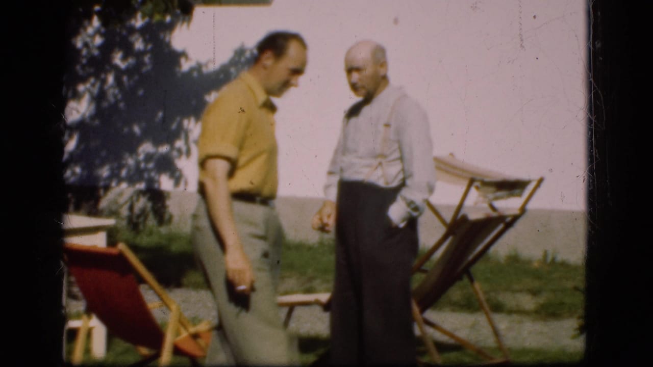 Two men stand near deck chairs on a lawn, with one man facing the other.