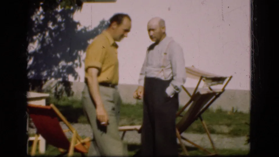 Two adult men in 1970s clothing stand beside garden chairs on a lawn in Denmark, with a white building behind them.