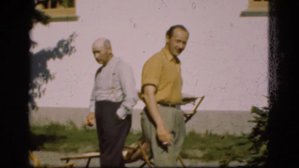 The two men move through the garden near a wooden chair, captured with soft handheld 8mm framing.