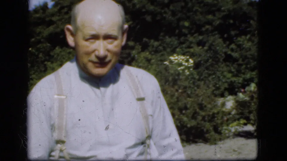 Older man faces the camera in a garden, wearing a pale shirt and suspenders, with hedge and flowers in the background.