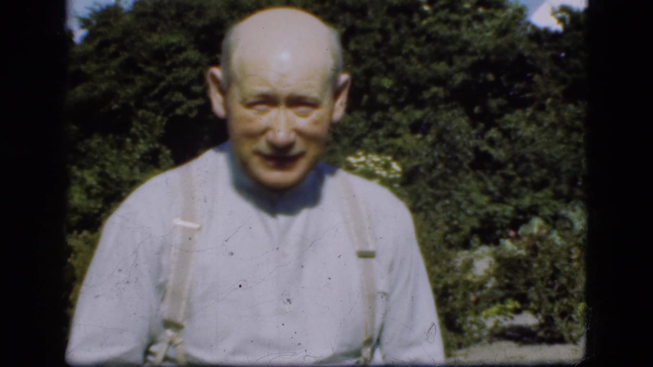 Older balding man in a light shirt and suspenders stands in a sunny garden.