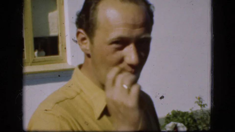 A younger man in a yellow collared shirt looks toward the camera near a window and pale exterior wall, with garden plants below.