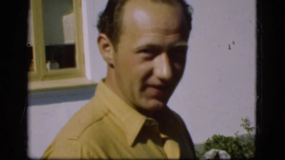 In Denmark circa 1975, a man in a yellow shirt is framed close-up outside a house in soft 8mm home-movie color.