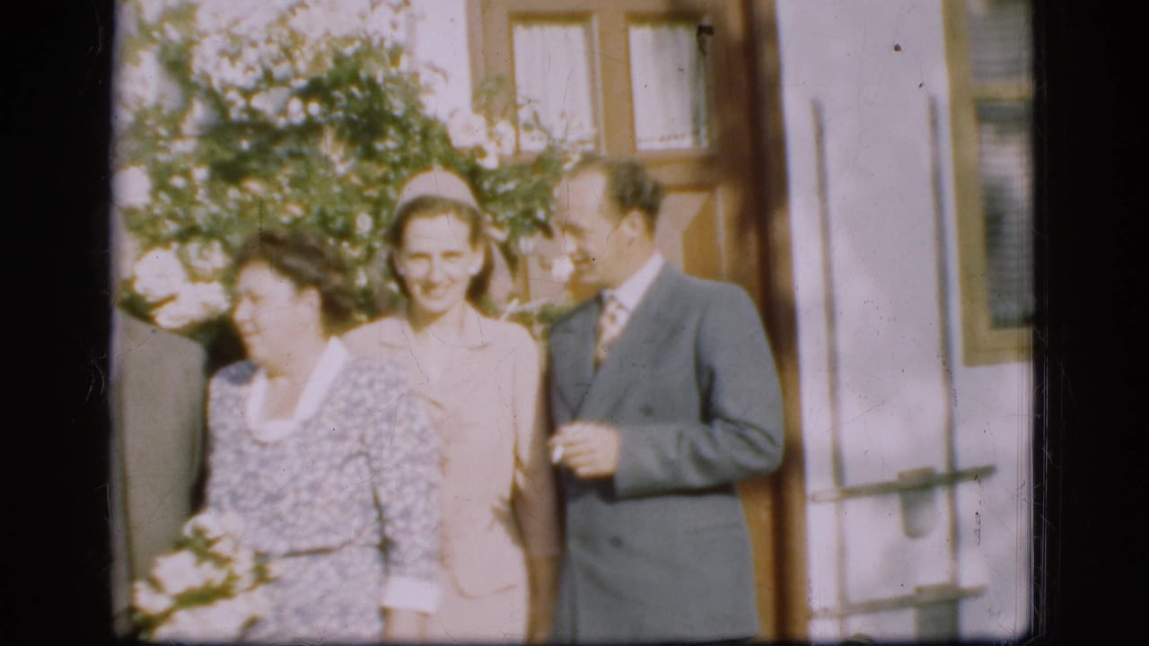 Adults in formal clothing stand outside a house near greenery and a doorway.