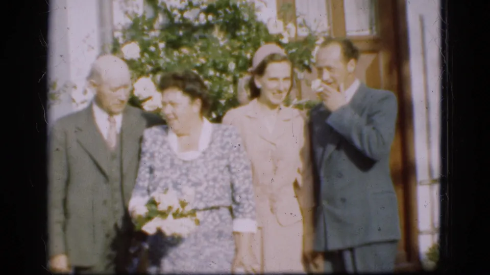 Four adults in formal clothing stand outside a Danish house in 1975, with flowers, greenery, and warm 8mm color visible.