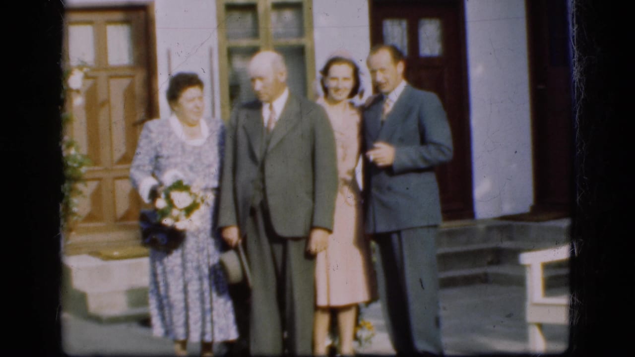 Four adults stand near building doors and steps, with a woman holding a bouquet.