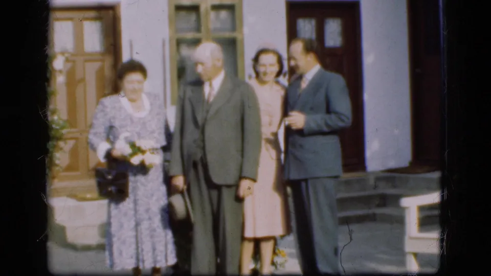 1975 Denmark home-movie frame of adults in formal attire outside a building, with the woman in front holding white flowers.