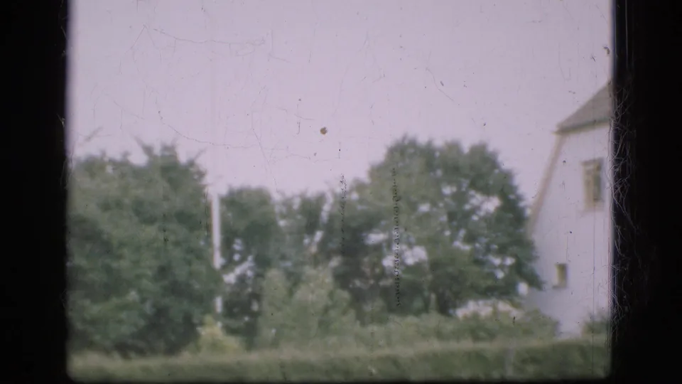 A Denmark 1970s 8mm frame shows trees and shrubs with a partial pale building at the edge of the view.