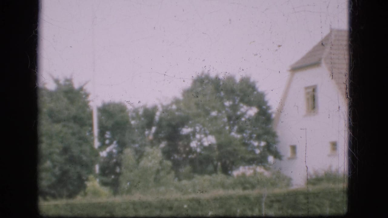 1975 Denmark 8mm frame showing a white farmhouse partly obscured by window framing, with trees, hedge, and pale sky.