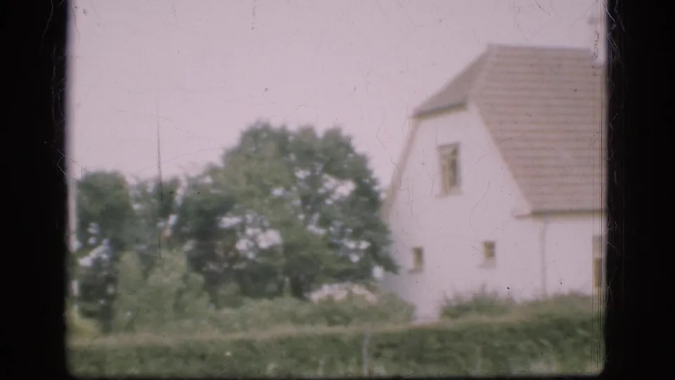 1975 Denmark rural view of a white farmhouse and tiled roof beyond hedge and trees, with 8mm grain and scratches visible.