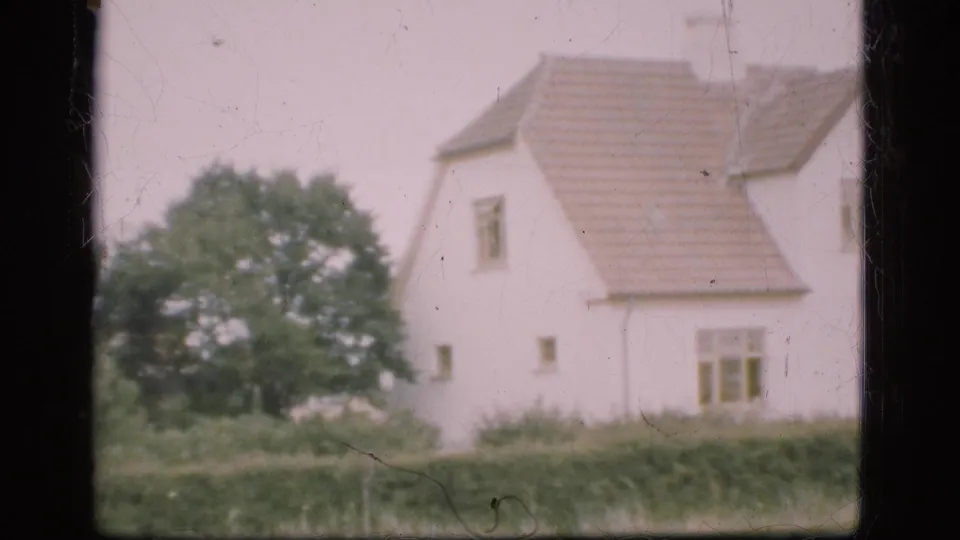Soft 8mm frame from Denmark in 1975 showing the farmhouse roof, windows, hedge, and trees as the pan continues right.