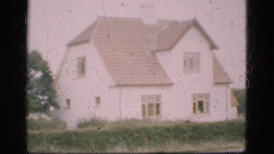 1975 Denmark 8mm frame showing the farmhouse more fully, including tiled roof, windows, hedge, trees, and visible film dust.