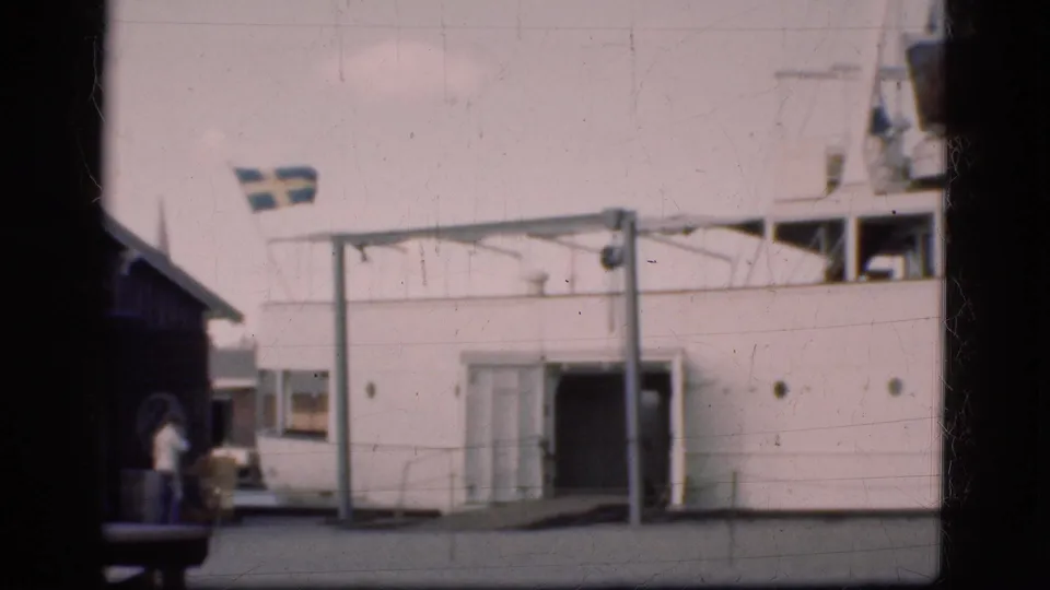 Home-movie frame of the ferry dock area in Denmark, with a Swedish flag above the vessel side and people standing near the boarding point.