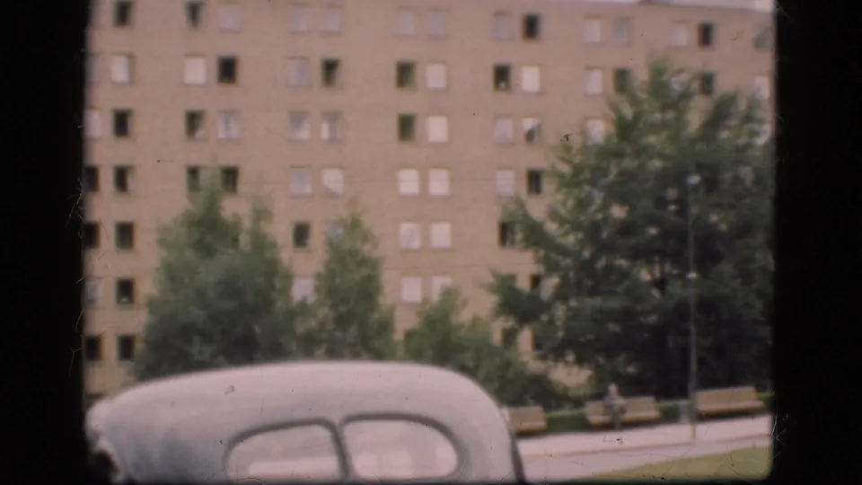 1975 Denmark street view with a rounded older car in the foreground, trees along the sidewalk, and a large apartment block behind.
