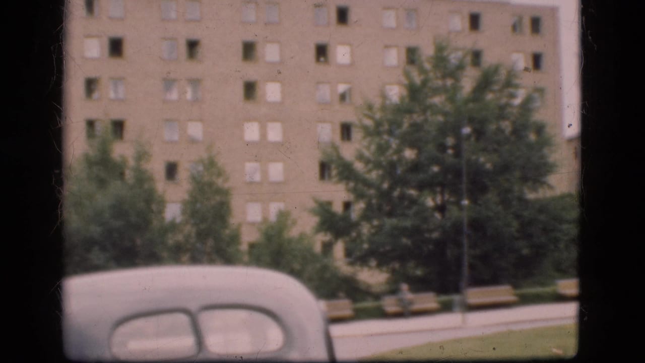 1975 Denmark home-movie frame showing the rear of an older light-colored car, park benches, trees, and a large multi-story building.