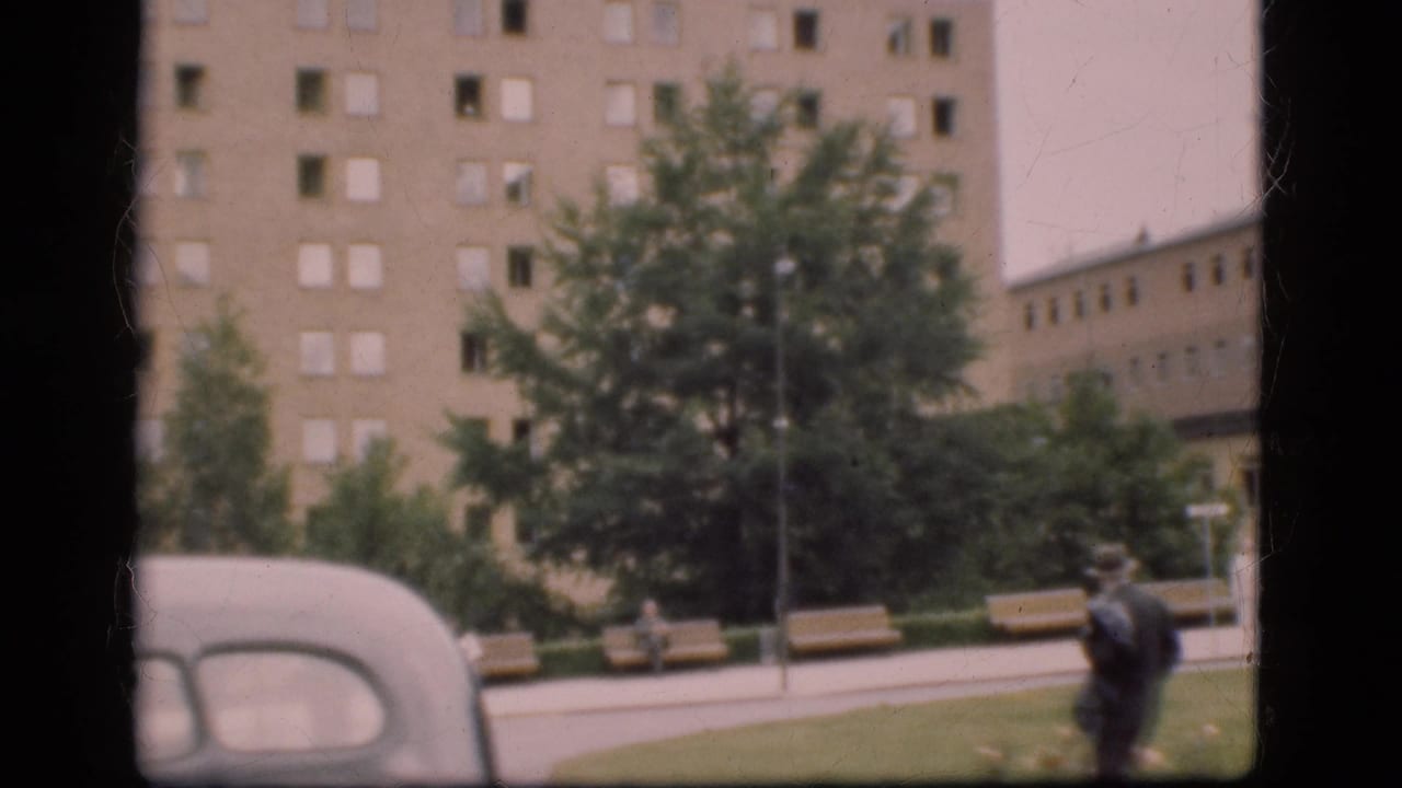 A man in a dark suit walks near benches and trees, with an older car and large building in view.