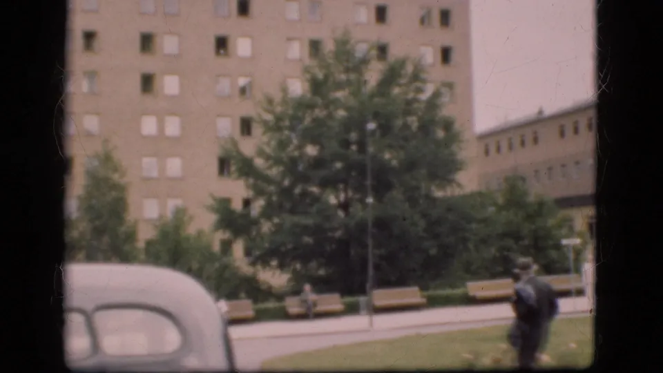 1975 Denmark frame with a suited man walking across a grassy urban space near benches, trees, an older car, and a large building.