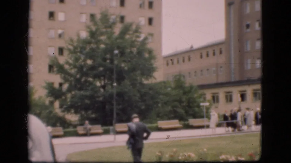 1975 Denmark home-movie frame showing a suited man crossing a lawn toward several people near benches and institutional-looking buildings.