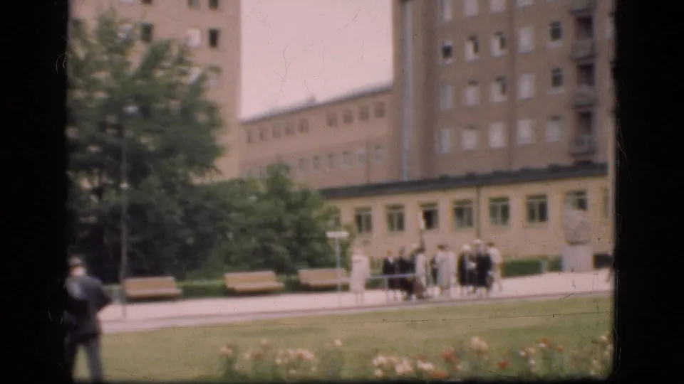 1975 Denmark frame with a suited man in the foreground, a small group near benches, trees, lawn, and large windowed buildings behind.