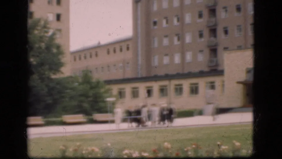 In Denmark in 1975, a small group of people walks near benches and lawn in front of a large modernist institutional-looking building.