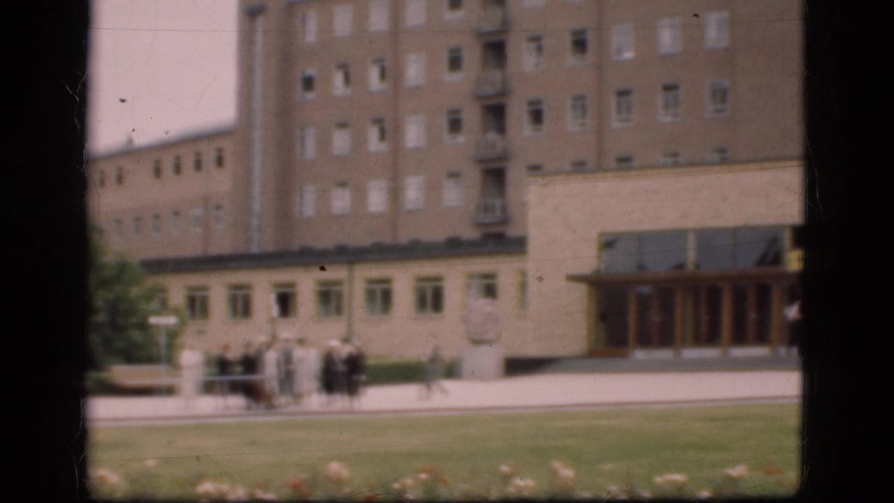 A distant 1975 Denmark view shows people near a broad paved area, lawn, and a large windowed building with an entrance section to the right.