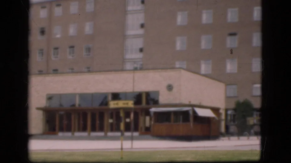 A soft 8mm frame shows a large Denmark building exterior in 1975 with an entrance, lawn, paved foreground, and a streetcar or tram-like vehicle near the front.