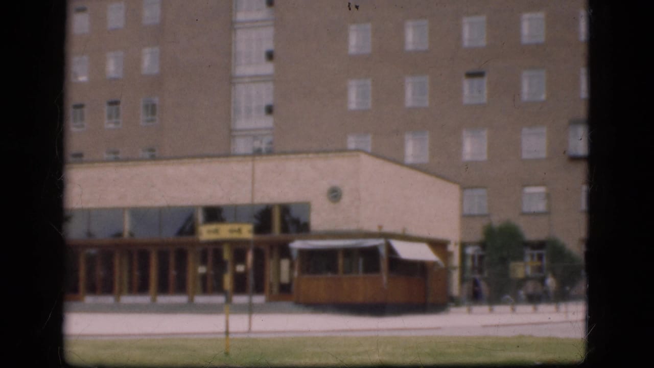 1975 Denmark 8mm frame showing a brick apartment block, storefront area, wooden kiosk, pedestrians, grass, and a road edge.