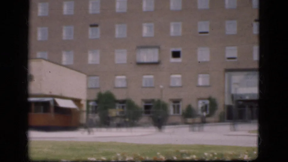 Soft 8mm Denmark frame showing the side of a kiosk, pedestrians crossing pavement, a brick building entrance, and a grass verge.