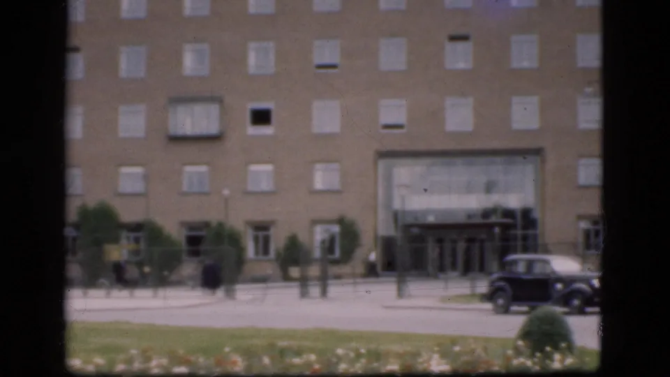 1975 Denmark frame showing a brick building entrance, pedestrians on the pavement, a signpost, flower bed, grass, and a dark rounded car.