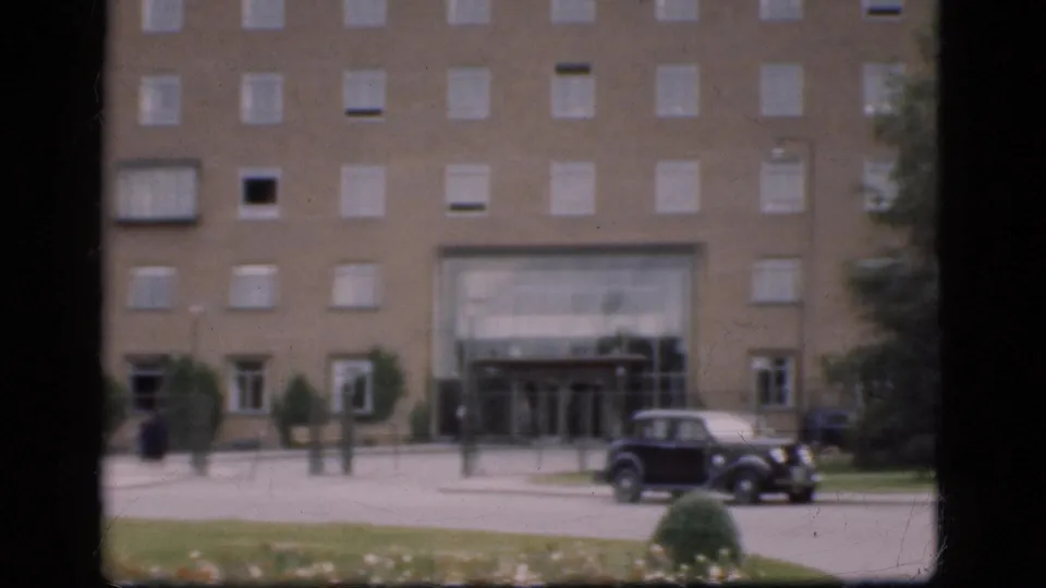 1975 Denmark 8mm frame showing a large building entrance, parked dark car, lawn, shrubs, and dark framing from a window or gate.