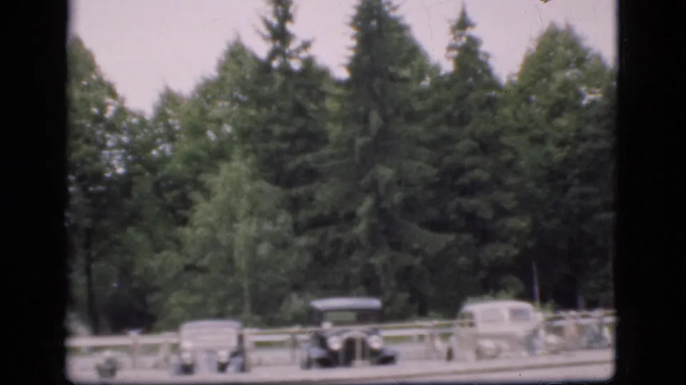 1975 Denmark 8mm frame with several classic cars parked near a pale railing, backed by dense evergreen trees.