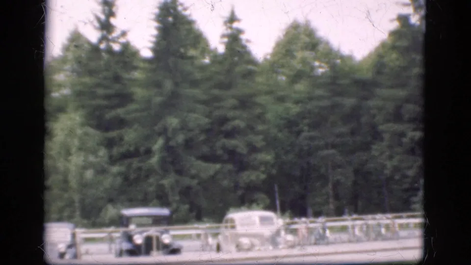 1975 Denmark 8mm frame showing classic cars, bicycles by a railing, paved roadway, and dense trees behind the parking area.