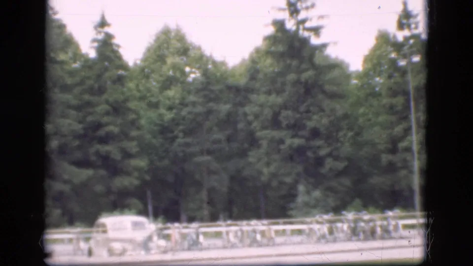 1975 Denmark 8mm frame with a light-colored classic car, bicycles, roadside railing, and leafy trees beyond.