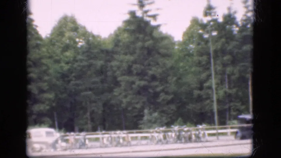 1975 Denmark 8mm frame showing a tree-lined road, a white car, roadside railing, and parked bicycles in soft focus.