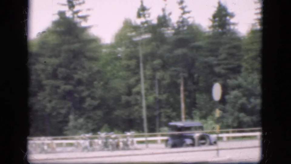 1975 Denmark 8mm frame showing a dark car by a roadside railing, dense trees, and a round sign.