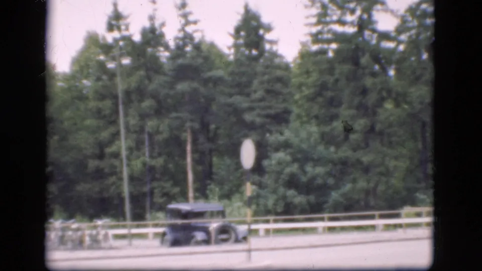 1975 Denmark 8mm frame showing a dark car passing a round traffic sign along a tree-lined road.