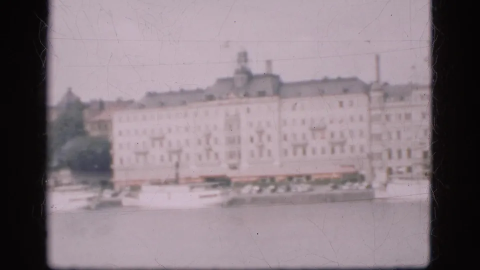 Soft 1975 Denmark 8mm view of a riverbank, pale multi-story buildings, docked boats, and dark home-movie frame edges.