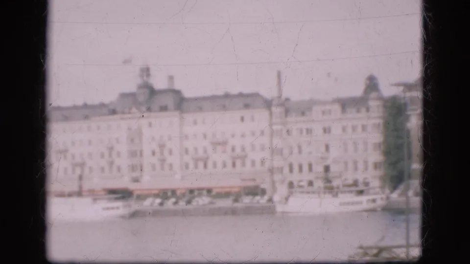 1975 Denmark 8mm frame showing a hazy waterfront panorama with pale buildings, moored boats, water, trees, and a distant dome.
