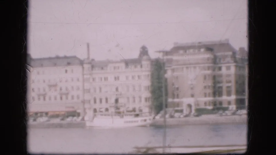1975 Denmark frame with large waterfront facades and a white boat near the shore, captured in faded 8mm color.